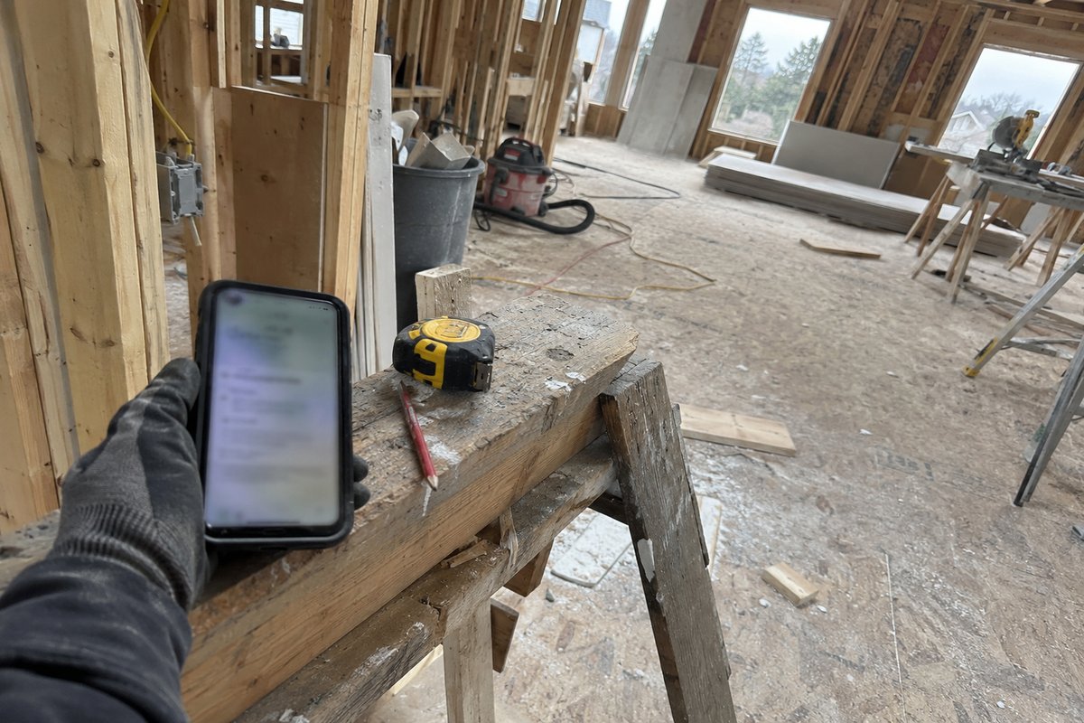 A gloved hand holding a phone over a sawhorse with a tape measure and pencil, inside a framed room under construction, flat daylight.