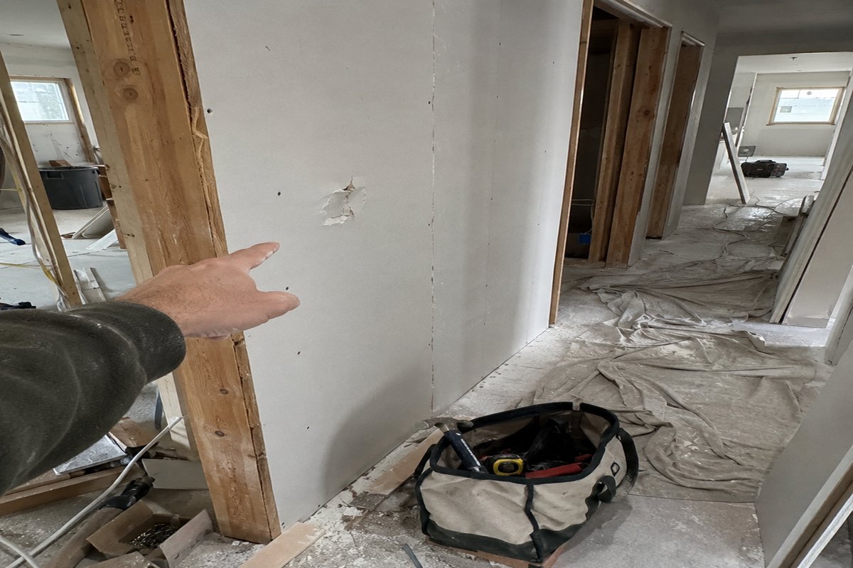 A hand pointing at a dented patch of drywall in a half-framed hallway, beside a half-packed tool bag on a drop cloth, flat daylight.